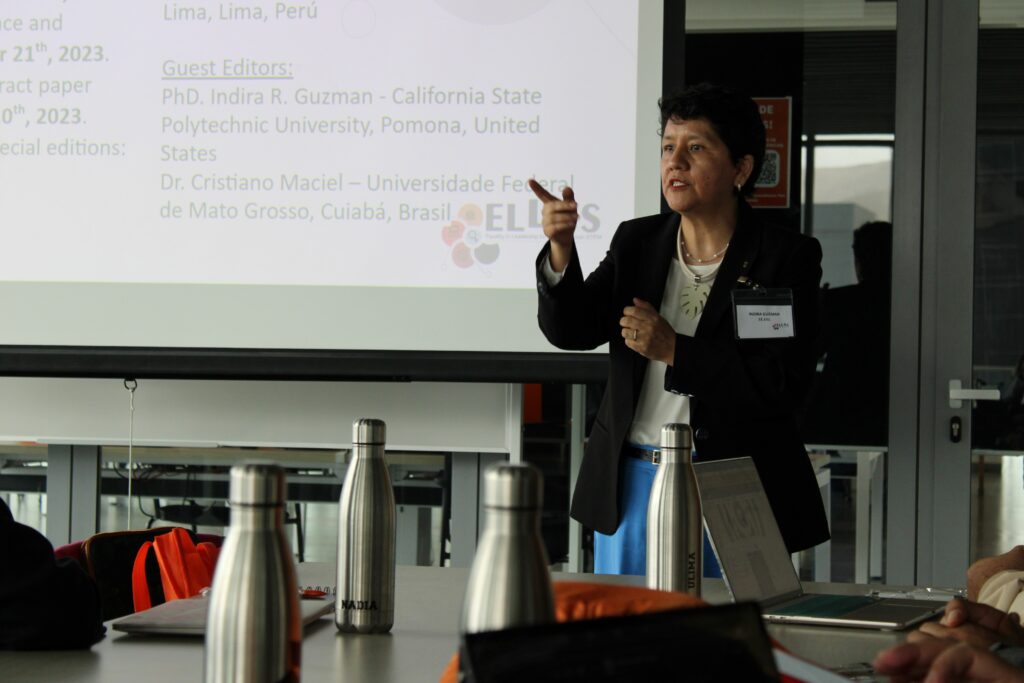 Foto de Indira Guzmann apresentando um trabalho do ELLAS durante o evento. Indira se encontra em uma sala, é possível ver um slide com informações da sua apresentação