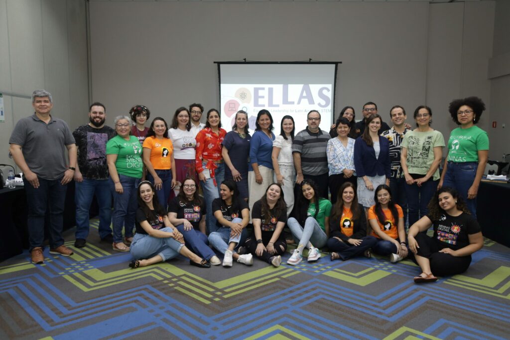 Membros do projeto posando para foto. Ao fundo é possível ver painel do ELLAS.